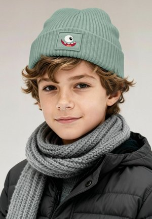 Light green ribbed knit beanie with a patch featuring a cartoon-like monster face; paired with a thick gray knitted scarf and a black jacket.