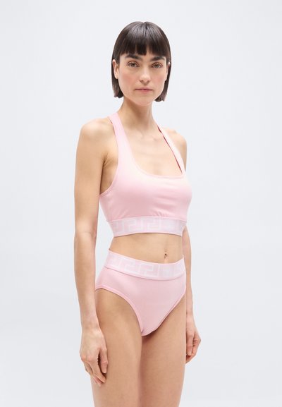 Γυναίκα με κοντά σκούρα μαλλιά που φοράει ανοιχτό ροζ αθλητικό μπουστάκι και αντίστοιχο εσώρουχο με ψηλή μέση, μπροστά σε απλό λευκό φόντο.