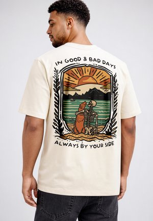 Man wearing beige shirt with sunset, mountain, dog, skeleton, and text "In good & bad days, always by your side" on back.