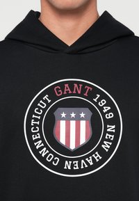 Fekete kapucnis pulóver, kör alakú grafikával, amelyen a "GANT 1949 NEW HAVEN CONNECTICUT" felirat és egy pajzs dizájn piros és fehér csíkokkal.