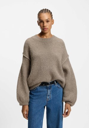 Chunky, alpaca-blend trui in lichtbruin, met een ronde halslijn en oversized mouwen. Gepaarde met hoge blauwe spijkerbroeken en een zwarte riem.