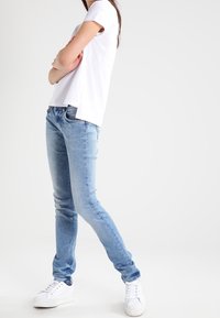 Světle modré denimové džíny s úzkým střihem, obohacené o klasický pětikapsový design a opotřebovaný vzhled, v kombinaci s bílými teniskami a bílým tričkem.