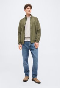 Fiatal férfi áll, olívazöld dzsekit, világosbézs pulóvert, kék farmert és világosbarna hasítottbőr csizmát visel, világosszürke háttér előtt.