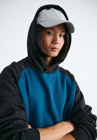 Felpa con cappuccio in tessuto teal e nero, dal taglio morbido, con maniche lunghe e polsini a costine, abbinata a un berretto grigio chiaro.