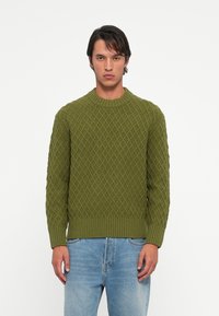 Pulover verde tricotat cu un model în diamant, decolteu rotund ribetat și tiv ribetat. Asortat cu blugi din denim bleu deschis.