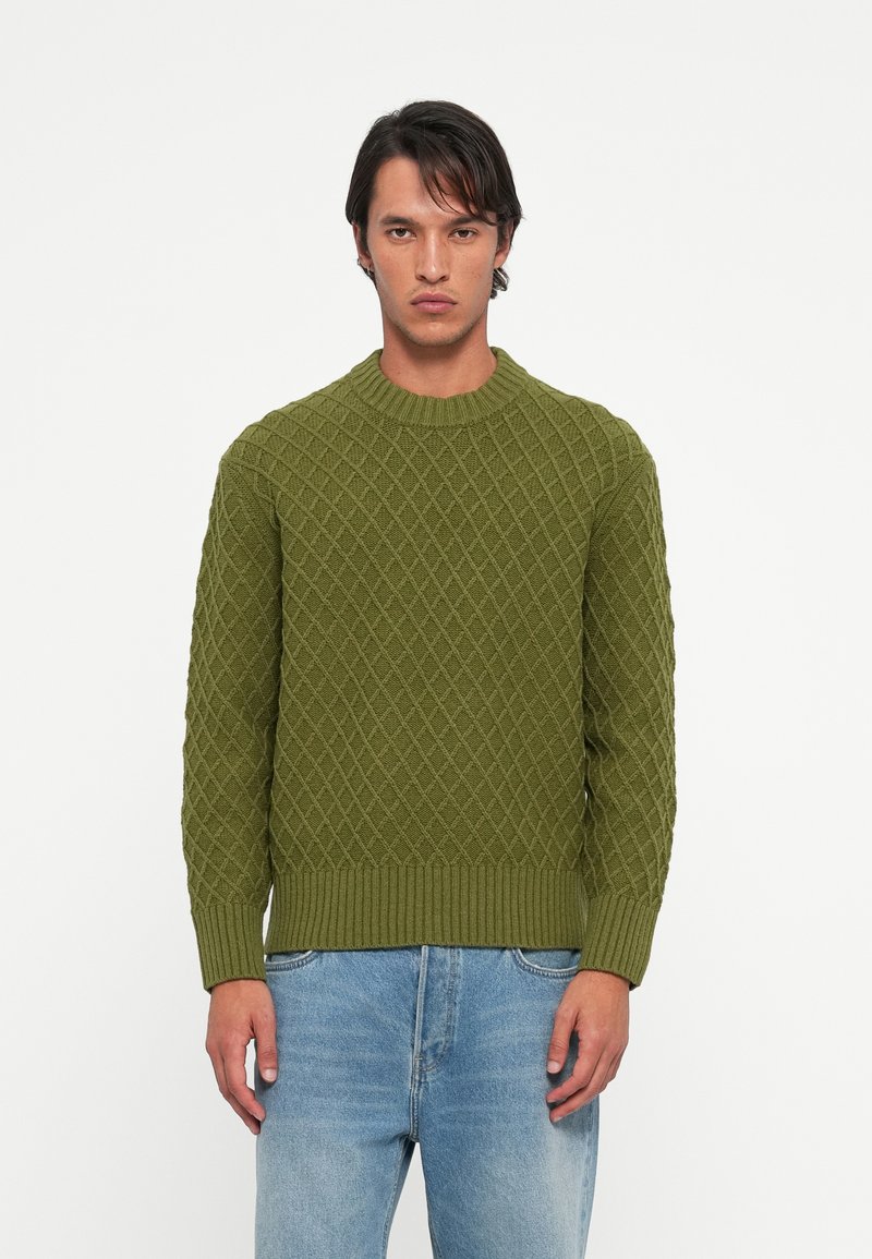 Pulover verde tricotat cu un model în diamant, decolteu rotund ribetat și tiv ribetat. Asortat cu blugi din denim bleu deschis.