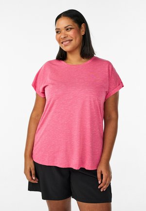 Smilende kvinde iført en lyserød T-shirt med korte ærmer og sorte shorts, stående foran en ensfarvet hvid baggrund.