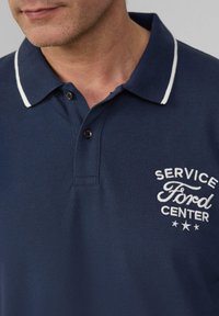 Man in een marineblauwe polo met witte bies op de kraag en 'Service Ford Center' in het wit geborduurd op de borst.