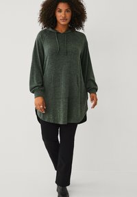 Vrouw met krullend haar, gekleed in een donkergroene hooded lange sweatshirt, zwarte broek en zwarte enkellaarzen, staande tegen een effen lichte achtergrond.