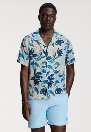 Camicia a maniche corte con bottoni caratterizzata da un motivo floreale blu, giallo e beige. Abbinata a pantaloni corti azzurri. Tessuto morbido.