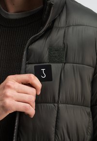 Dunkelgraue Pufferjacke mit matter Oberfläche. Verfügt über ein grünes Stoff-Patch und ein schwarzes Marken-Patch mit weißem Logo. Strukturiertes Strickmaterial darunter.