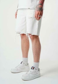 Shorts di denim bianchi con orli sfrangiati e dettagli strappati, abbinati a sneakers bianche e calze grigie a costine. Le gambe sono leggermente abbronzate.