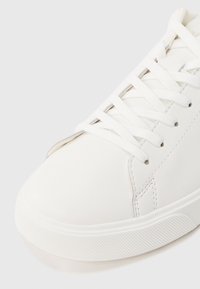 Κοντινή λήψη λευκού αθλητικού παπουτσιού με λευκά κορδόνια και υφασμένο λαστιχένιο πέλμα σε απλό ανοιχτό φόντο.