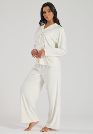 Two-piece pajama set in soft, cream-colored fabric. The top features a collar, buttons, and a chest pocket; the pants have a relaxed fit.