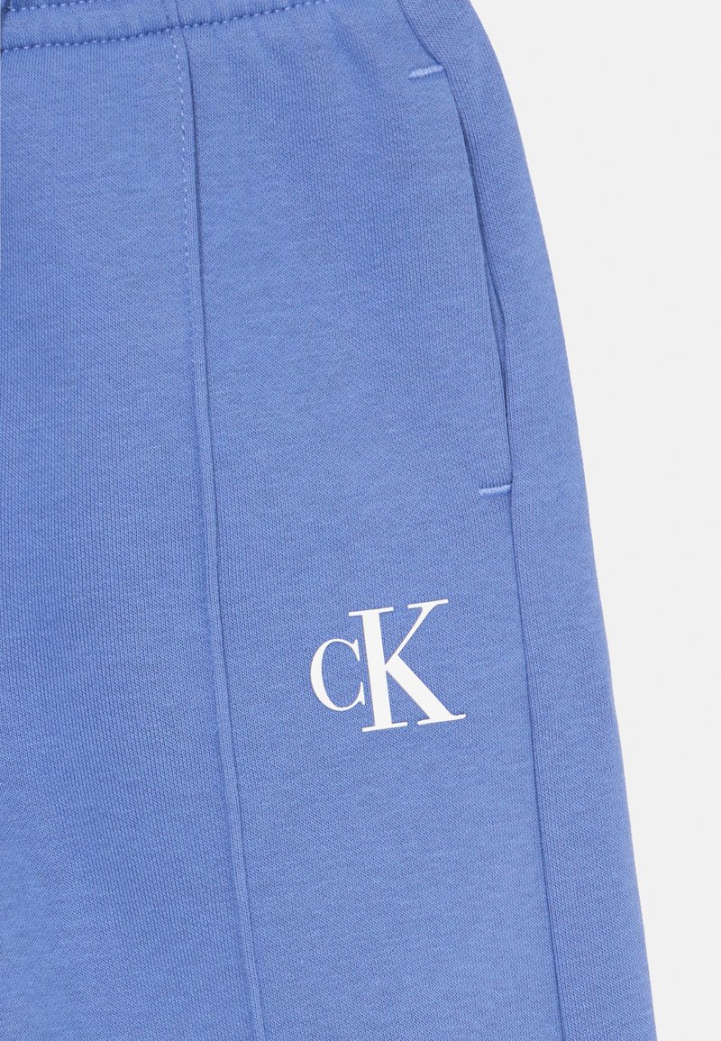Primo piano di un tessuto blu che mostra un logo bianco "cK", una cucitura laterale e una tasca nascosta laterale su un indumento.