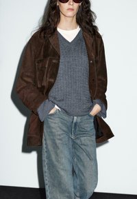 Persona che indossa una giacca scamosciata marrone scuro, un maglione grigio a trecce sopra una camicia bianca, jeans blu larghi, mani in tasca e occhiali da sole grandi.