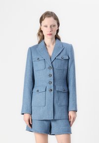 Kék tweed blézer két elülső zsebbel, hat gombbal és strukturált gallérral, amelyet hozzáillő sorttal párosítottak a harmonikus megjelenés érdekében.