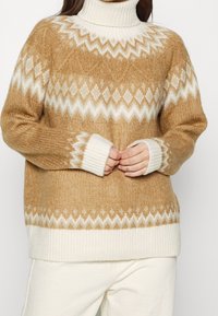 Stickad tröja i beige och krämfärg, med hög krage, geometriska mönster och ribbade muddar. Mjuk textur med en kraftig design.