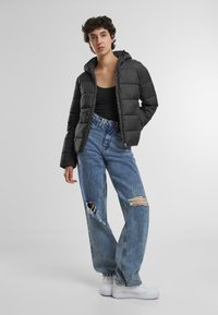 Svart puffjakke med hette, med kviltet design. Kombinert med høytlivs, slitte lyseblå jeans og hvite joggesko.