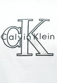 Biela bavlnená tkanina so vzorom vyšitého textu „Calvin Klein“ a veľkým logom „CK“ v tmavošedej farbe. Hladká textúra s čistými líniami.