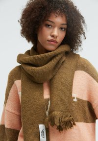 Young woman with curly hair wearing a brown knit scarf and a pink and brown striped sweater, looking directly at the camera.