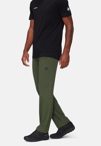 Green softshell pants with a slim fit, side pockets, and a black logo. Paired with a black short-sleeve shirt and black footwear.