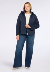 Jonge vrouw in een marineblauwe gewatteerde rits-hoodie, wit topje, wijdvallende blauwe jeans en witte sneakers, staande tegen een effen achtergrond.