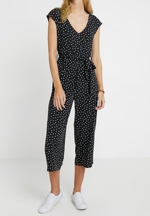 Czarny kombinezon w białe kropki, dekolt w serek, krótkie rękawki, wiązany w pasie, spodnie o długości do połowy łydki, lekki materiał, w zestawieniu z białymi trampkami.