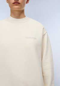 Crème-kleurige sweatshirt met een ribbelstructuur bij de ronde hals en verlaagde schouders. Heeft een subtiel geborduurd logo op de voorkant. Zachte stoftextuur.