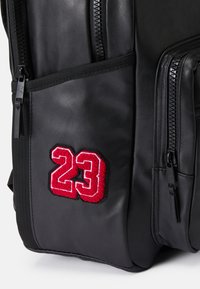 Mochila sintética preta com superfície texturizada, apresentando dois fechos de correr e um "23" bordado em vermelho e branco de um dos lados.