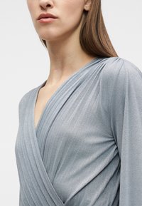 Γυναίκα που φοράει ένα λαμπερό ασημί φόρεμα τύπου κιμονό με πλισέ λεπτομέρειες στον ώμο και το μπούστο, μπροστά σε ένα απλό φόντο.