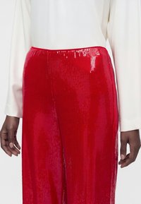 Personne portant un haut blanc à manches longues et un pantalon rouge à paillettes, taille haute et coupe ample, montrée de la taille au milieu de la cuisse.