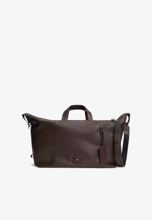Ciemnobrązowa skórzana torba podróżna typu duffel z podwójnymi uchwytami, odpinanym czarnym paskiem na ramię oraz małą prostokątną naszywką z logo z przodu.