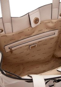 Beige draagtas met een textuur op de buitenkant, voorzien van gouden hardware en een binnenzak. De binnenvoering heeft een herhalend "GUESS" logo-patroon.