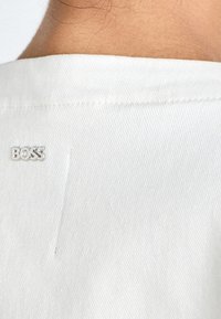 Valkoinen farkkupaita, jossa on näkyvä sauma ja hopeinen "BOSS"-logo yläselässä, tuoden brändin lisäyksen muotoiluun.