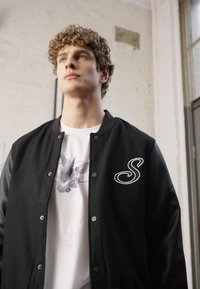 Sort varsity-jakke med læderærmer, der har et hvidt broderet 'S' logo. Underneath en hvid t-shirt med et blomstergrafisk design.