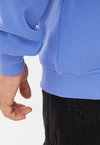 Lichtblauwe sweatshirt met ribgebreide manchet en zoom, gladde textuur, met een klein geborduurd logo dichtbij de heup, gecombineerd met donkere broek.