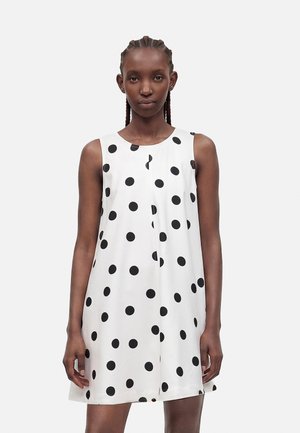 Donna con capelli intrecciati che indossa un vestito bianco senza maniche con grandi pois neri, in piedi davanti a uno sfondo chiaro e uniforme.
