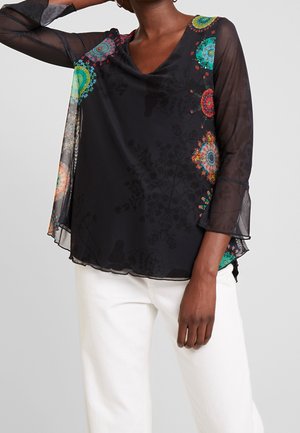 Femme porte un chemisier noir transparent à manches longues avec des motifs circulaires colorés, associé à un pantalon blanc, sur un fond gris uni.