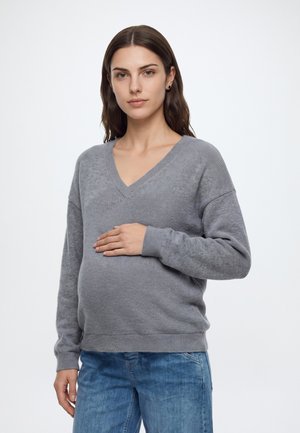 Femme enceinte en pull gris et jean bleu tenant doucement son ventre, debout devant un fond blanc uni.