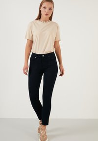 Camiseta beige con cuello redondo y letras negras, combinada con jeans ajustados de color azul oscuro que tienen bolsillos frontales y un cierre de botón visible.