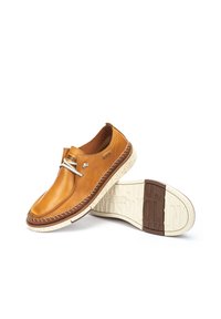 Zapatos de cuero color miel con un diseño redondeado, que presentan un ojal plateado, una suela marrón contrastante y una entresuela blanca moteada. Detalle de costura de acento.