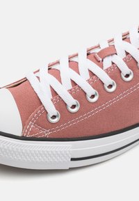 Zapatilla de lona rosa con cordones blancos, punta de goma y suela a rayas negras. Presenta ojales plateados y superficie superior texturizada.