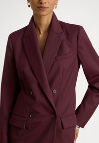 Μπορντό σακάκι διπλής σειράς με υφή, γιακάδες με σχισμές, δύο μπροστινές τσέπες και μαύρα κουμπιά.