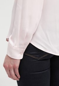 Blouse rose pâle avec une texture douce et transparente, manches avec poignets boutonnés et coupe décontractée, associée à un jean en denim foncé.