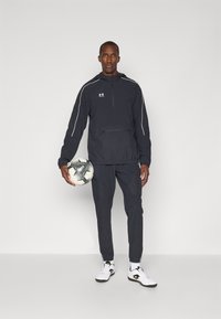 Black athletic jacket with white accents, half-zip, and kangaroo pocket, paired with matching pants and white shoes, holding a black and white soccer ball.