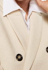 Blazer beige su misura realizzato in un tessuto morbido, con un classico design doppiopetto, grandi bottoni scuri e un colletto bianco sotto.