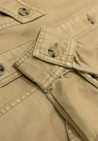 Beżowa tkanina z przeszyciami i zapięciami na guziki. Tekstura jest gładka z delikatnymi fałdami, podkreślając miękkość materiału.
