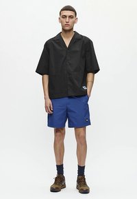 Junger Mann steht da, trägt ein lockeres schwarzes kurzärmliges Hemd mit Knöpfen, blaue Shorts, dunkle Socken und braune Wanderschuhe, vor einem einfarbigen Hintergrund.