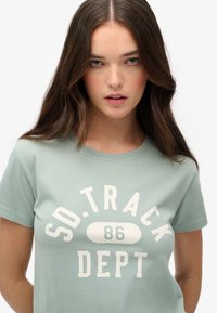 Lys grøn bomulds t-shirt med korte ærmer, der har et centralt hvidt grafik med teksten "SO. TRACK DEPT 86" i fed skrift.